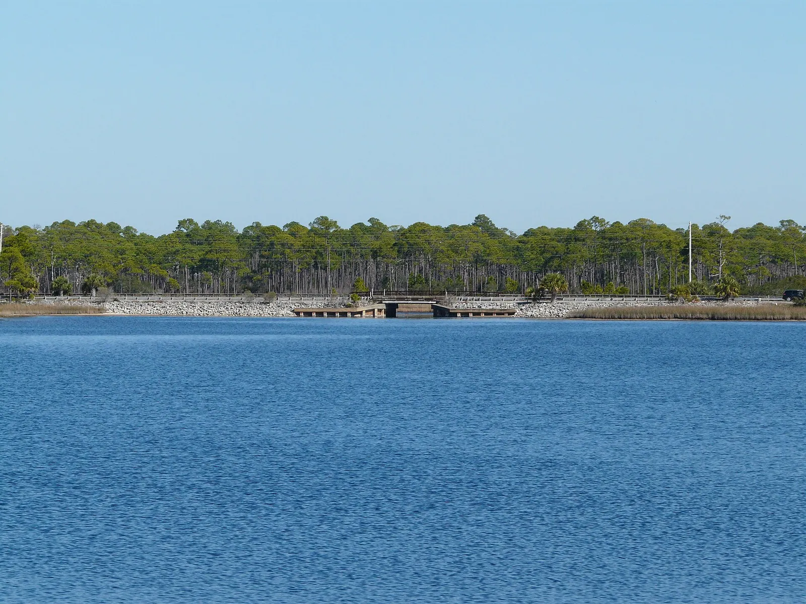 Calm blue Western Lake with the low County Road 30A bridge and pine forest along the far shore