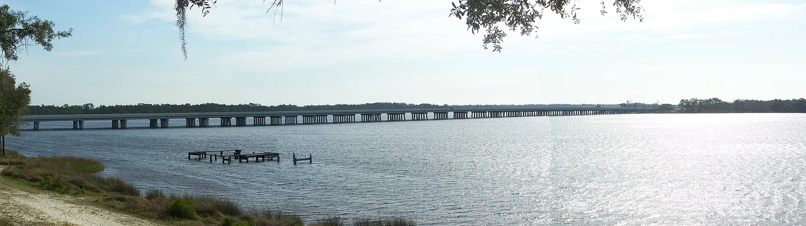 Wide view of Lake Powell’s calm water with a long highway bridge on the horizon, seen from the shore at Camp Helen State Park