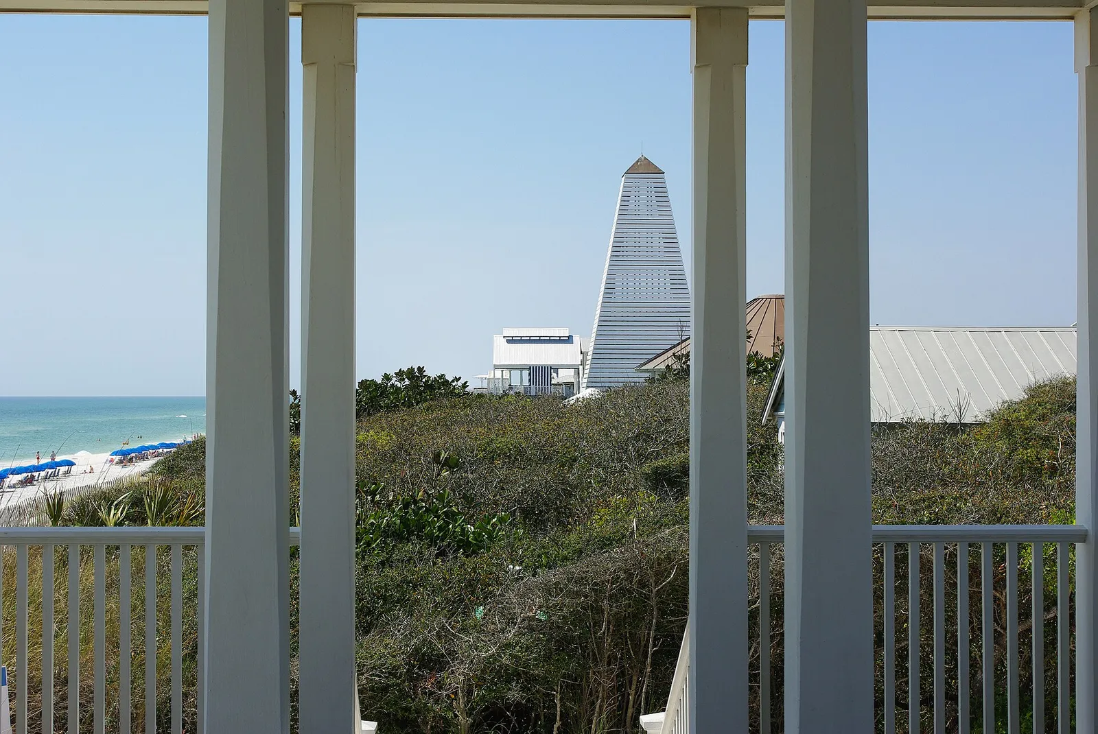 View between white porch columns of a Seaside beach pavilion toward the Gulf, beach umbrellas and a tall pyramid-roofed pavilion tower