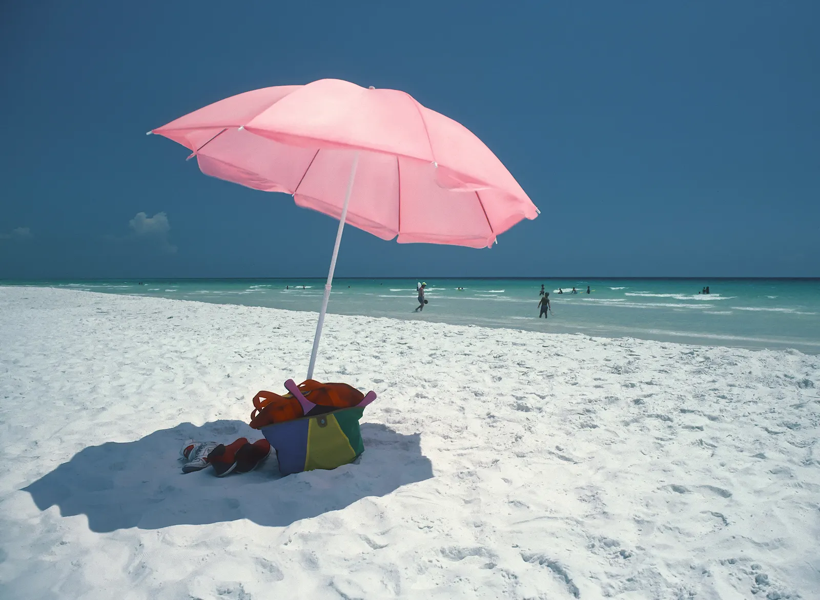 Pink beach umbrella and beach bag on bright white sand with swimmers in turquoise water at Seaside