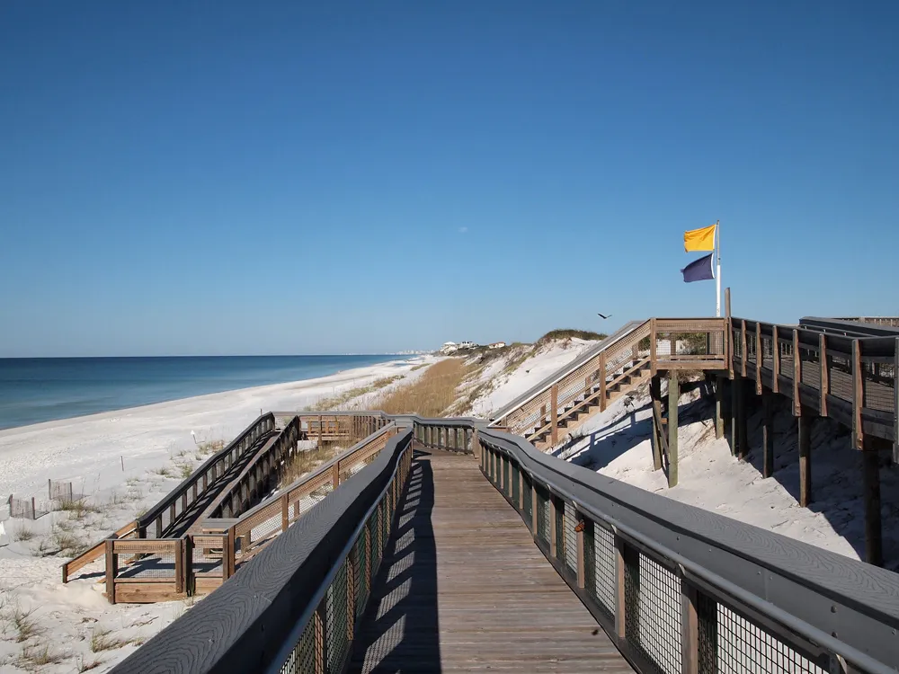 Wooden boardwalk and stairs crossing tall white dunes down to the beach at Grayton Beach State Park, beach flags flying